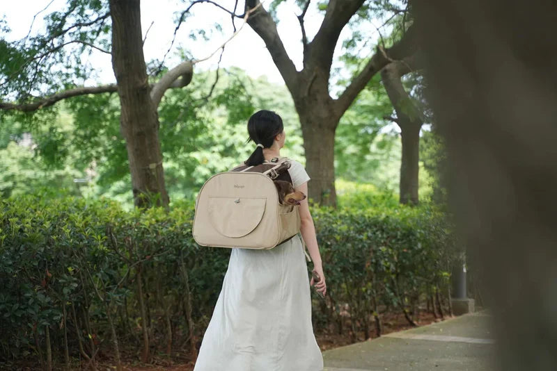 Woman walking outdoors with a beige backpack, surrounded by greenery.