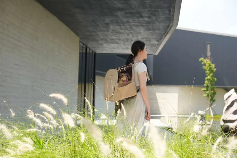Woman walking outdoors with a backpack, surrounded by greenery and a building.